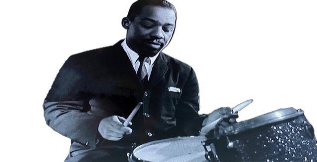 A young Art Blakey sitting at drums facing forward with eyes close.