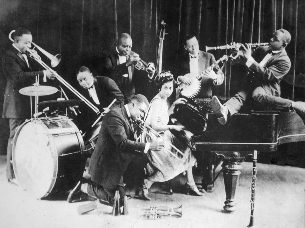 A black and white photograph of King Oliver's Creole Orchestra in 1923 with Louis Armstrong kneeling dead center with other band members behind him and to either side anti sly playing their instruments.