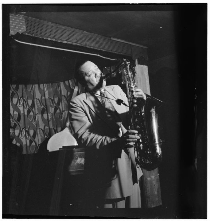 A black and white photograph by Robert P. Gottlieb of Lester Young standing and playing his saxophone at the Famous Door in New York City in September, 1946 in front of a geometric patterned curtain and a glimpse of a piano player's head in the background on the far left.