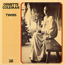 The album cover for "Twins" by Ornette Coleman with a bald women in a long tubular robe extending from neck to floor seated on a long wooden bench in a  columned interior courtyard with potted plants and a potted tree having a light sepia yellowed filter over entire photograph.