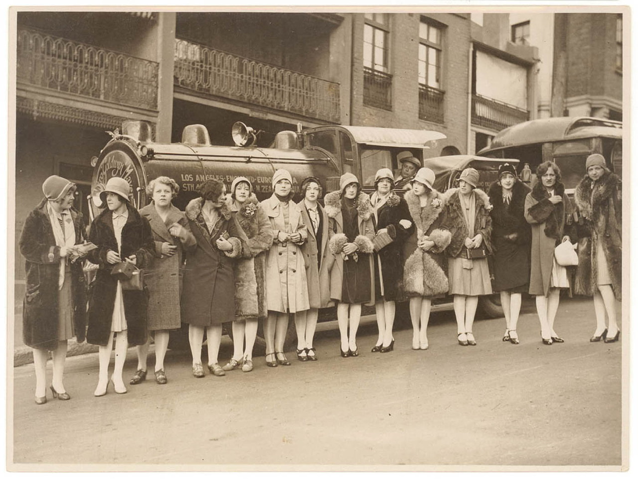 A sepia photograph of the all-woman band, The Ingenues, standing spread out in front of the first trackless train a Metro Goldwyn Meyer promotional vehicle for the movie "Ben Hur," in Sydney, Australia, ca. 1928.