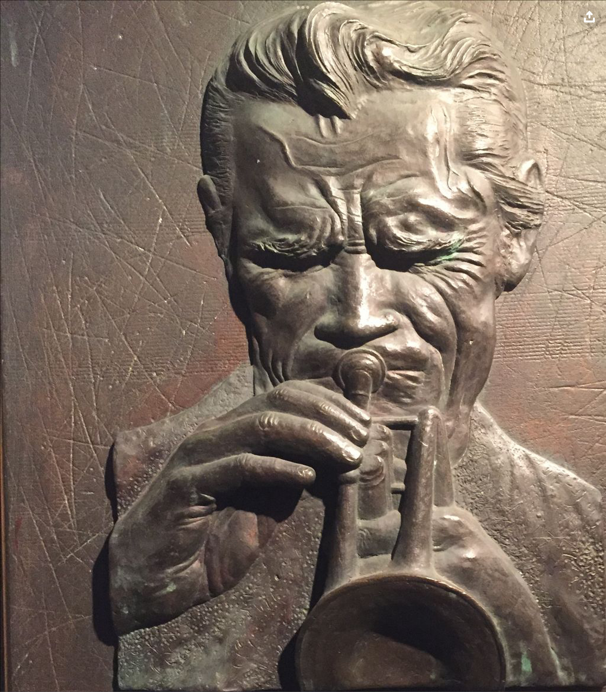 A closeup of a bronze plaque of Chet Baker with bushy eyebrows and eyes closed blowing furiously into his trumpet memorializing the location of Chet Baker's death at the Prins Hendrik hotel in Amsterdam with the writing at bottom of plaque cutoff.