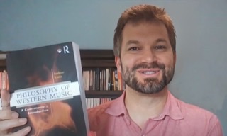 A color photograph of Andrew Kania smiling and holding up a copy of his book Philosophy of Western Music.