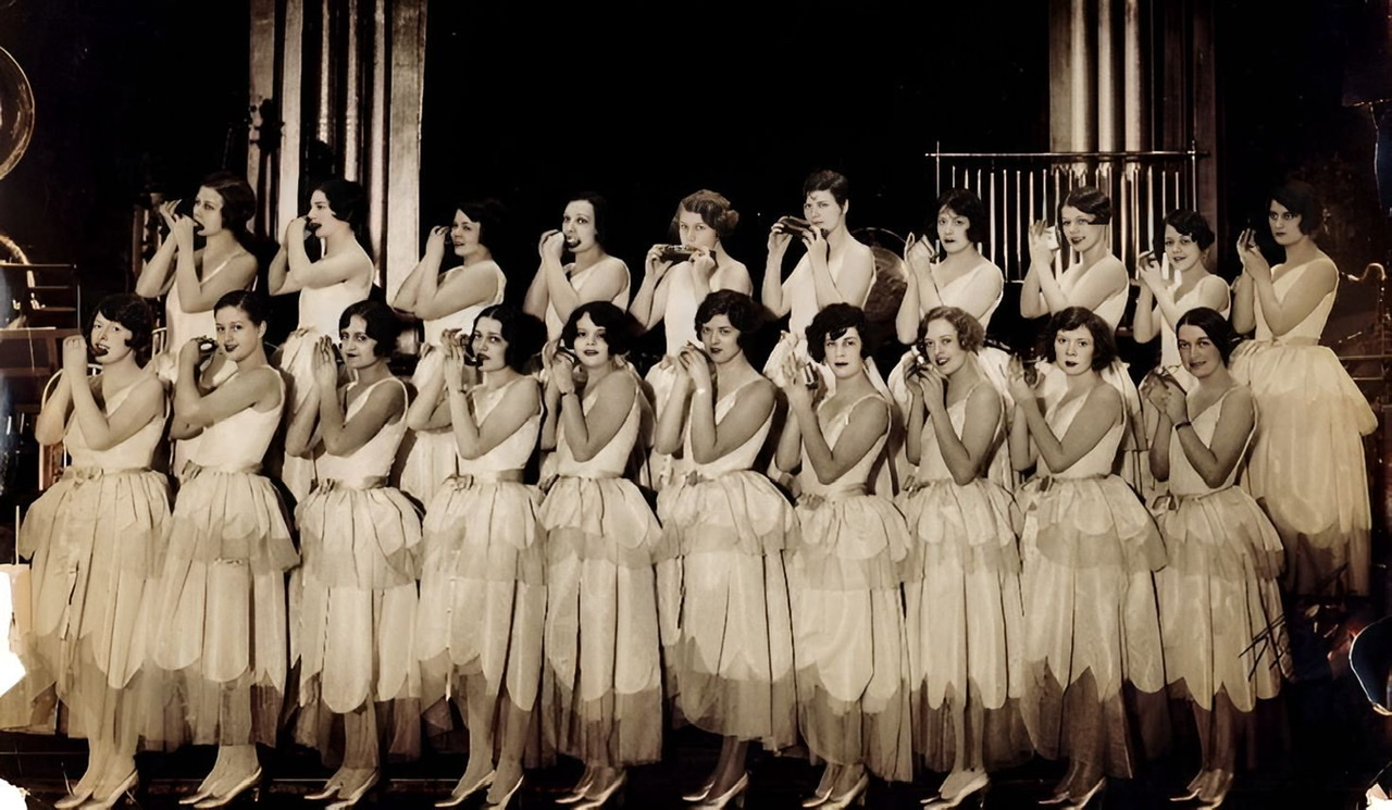 An enhanced photograph of twenty of The Ingenues lined across the stage in two rows each playing an harmonica of various sizes.