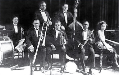 A different black and white photograph with five musicians seated including Louis Armstrong in center with King Oliver and William Johnson standing behind them in background all facing the camera with Lil Harden (Armstrong) seated at the piano on the right.
