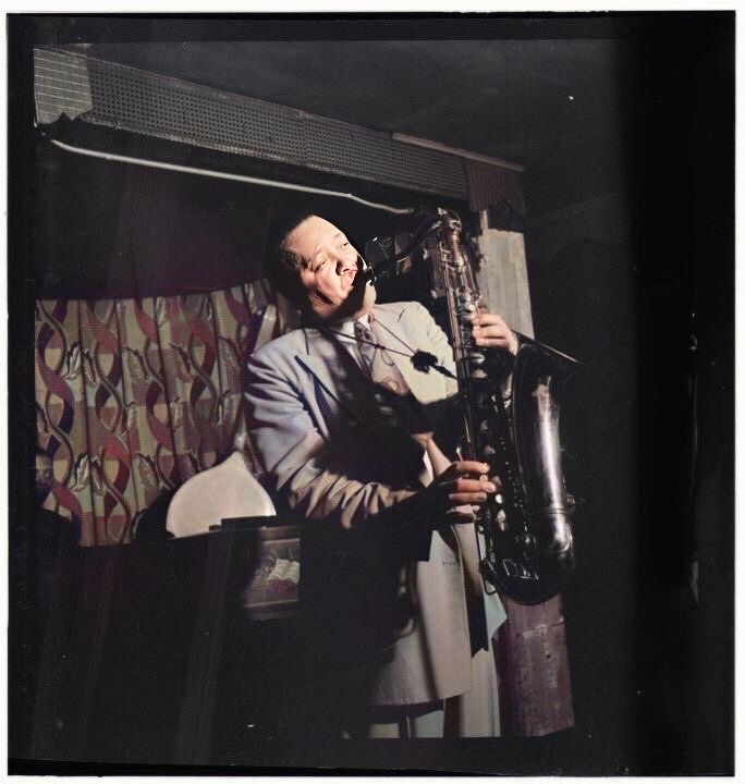 A colorized photograph by Robert P. Gottlieb of Lester Young standing and playing his saxophone at the Famous Door in New York City in September, 1946 in front of a geometric patterned curtain and a glimpse of a piano player's head in the background on the far left.