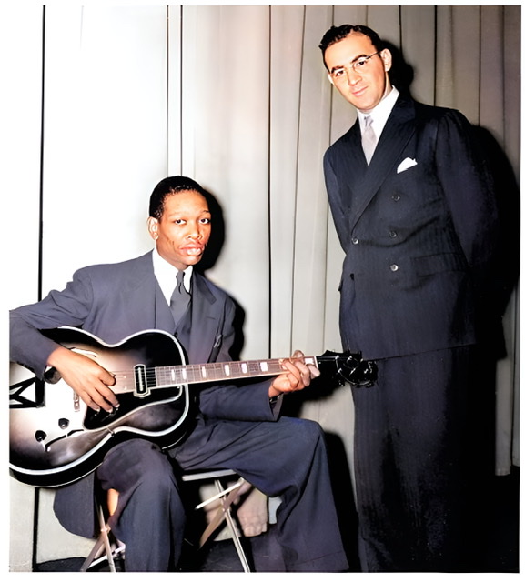 A colorized photograph Charlie Christian sitting in a chair holding his guitar in playing position with Benny Goodman standing on the right with both men wearing suits.