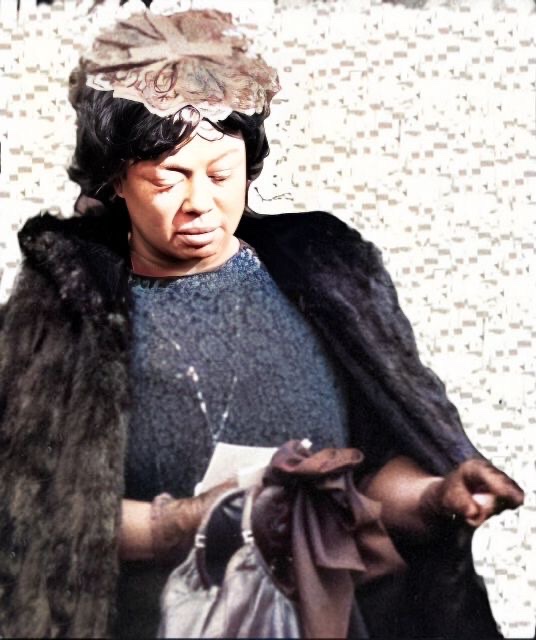 A colorized black and white photograph of a heavyset Hazel Scott in late middle age wearing a fur coat and carrying a leather double handle handbag.