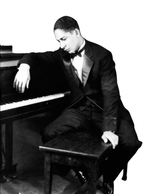 Jelly Roll Morton as a young man in a tuxedo sitting on a piano bench draped over the front of a piano with head bowed.