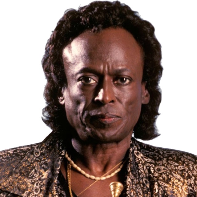 Miles Davis looking straight ahead with golden shirt and white beaded necklace.