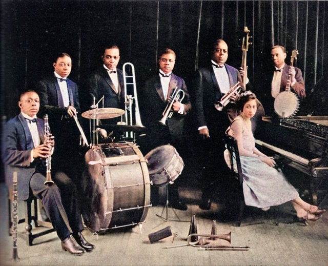 A colorized black and white photograph with Louis Armstrong and King Oliver centered with Lil Harden (Armstrong) seated at the piano on the right while other musicians surround them.