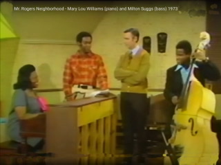 A screen capture of Mary Lou Williams seated playing piano on left with Mr. Rogers standing in middle and bassist Milton Suggs playing bass on right.