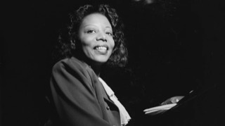 A William P. Gottlieb black and white photograph of Mary Lou Williams smiling sitting on a piano bench closeup of her from the waist up facing right and looking at camera 📸.