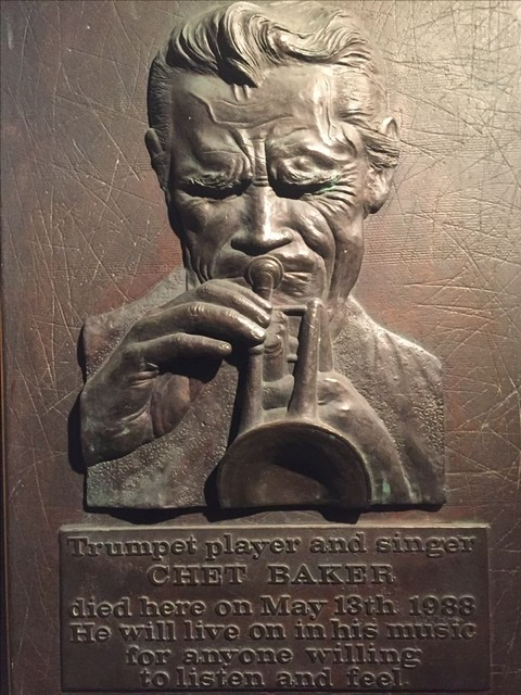 A closeup of a bronze plaque of Chet Baker with bushy eyebrows and eyes closed blowing furiously into his trumpet memorializing the location of Chet Baker's death at the Prins Hendrik hotel in Amsterdam with the writing at bottom showing the words "Trumpet player and singer Chet Baker died here May 13, 1988.  He will live on in his music for anyone willing to listen and feel."