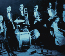 A black and white photograph with musician's names labeled by each one of King Oliver's Creole Orchestra in 1923 with King Oliver centered in the background and Louis Armstrong playing trumpet and at far right Lil Harden (Armstrong) seated at the piano.
