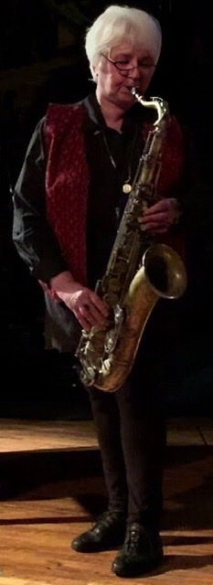 A detail of a color photograph of Lena Bloch standing and wearing a black ensemble with a red vest and black sneakers 👟while playing her saxophone.