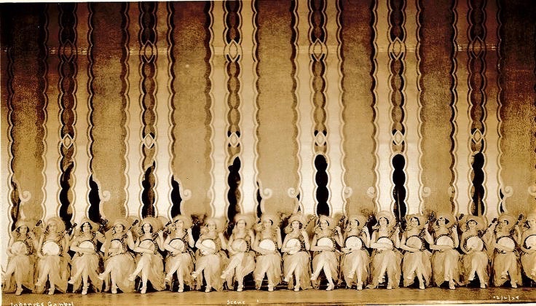 An enhanced sepia style photograph of nineteen of The Ingenues lined up on chairs across the entire stage each holding a banjo with crossed legs with a large multiply striped art deco curtain rising way up behind them.