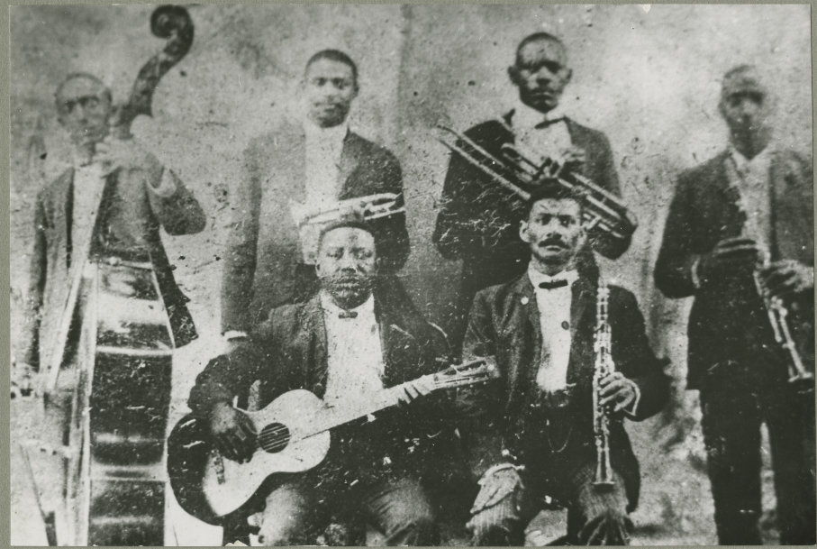 A black and white photograph of the Buddy Bolden band with Bolden second fron left holding his trumpet at his waist.