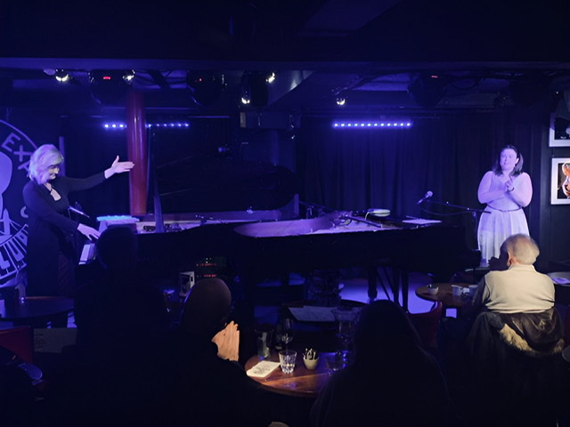 A color photograph of Dena DeRise on far left pointing to her co-pianist Champian Fulton on right with two Steinway pianos sandwiched between them.
