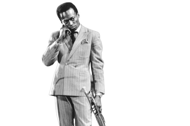 Performing onstage in West Germany around 1959 in a pin-striped suit with right hand on his right cheek with trumpet hanging down on his left side.